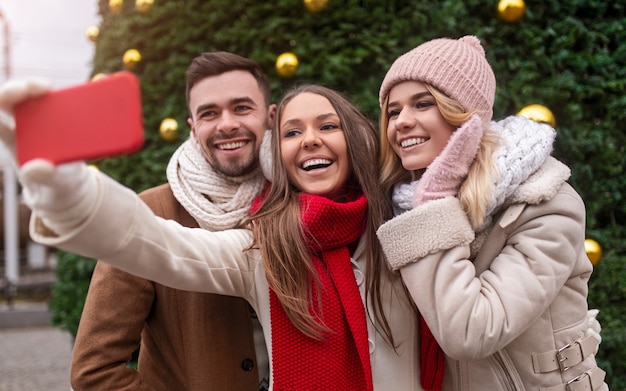 Grupa zachwyconych młodych przyjaciół w ciepłych ubraniach stojących na tle zielonej choinki i robiącej selfie na smartfonie podczas wspólnego spędzania wakacji w mieście