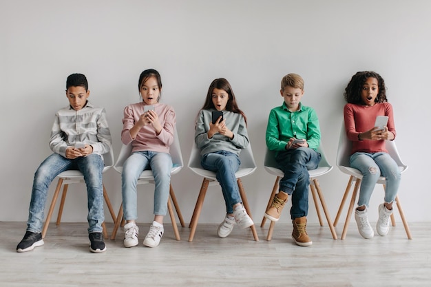 Grupa zdumionych, zróżnicowanych dzieci w wieku szkolnym korzystających ze smartfonów siedzących w pomieszczeniu