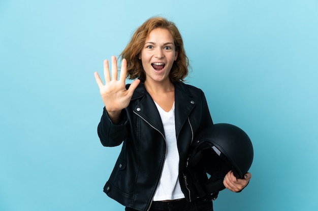 Gruzińska dziewczyna trzyma kask motocyklowy na białym tle na niebieskiej ścianie, licząc pięć palcami
