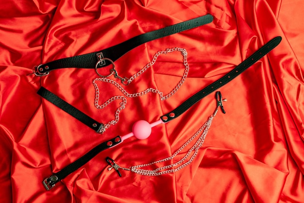 Gry erotyczne dla dorosłych. Elementy BDSM. Skórzany obroża z metalową łańcuszkową smyczą, kulka z różowym kneblem, zaciski na sutki na czerwonej satynowej prześcieradle.