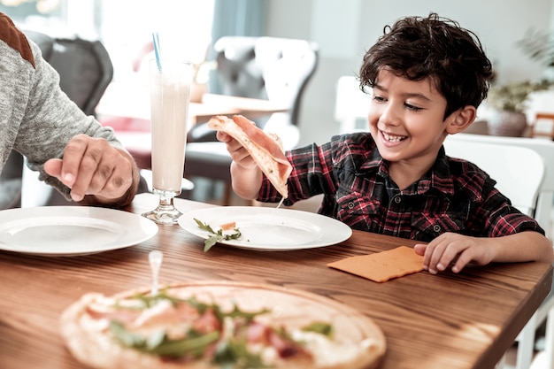 Gryząca pizza. Emocjonalny zabawny ojciec gryzący kawałek pizzy z bekonem i zielenią z rąk syna