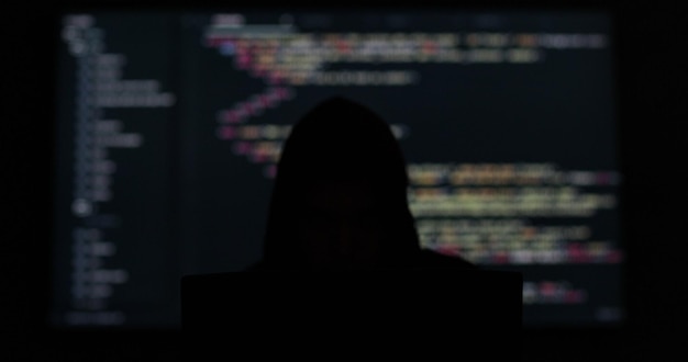 Hacker w kapturze pracujący w ciemnym, abstrakcyjnym pokoju, kod programowania na dużym ekranie w tle, bezpieczeństwo systemu cyfrowego.
