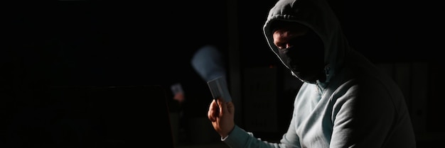 Haker w ciemnej kurtce patrzący na ekran laptopa w ciemnym pokoju trzyma kartę bankową
