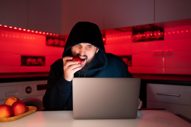Haker w czarnej bluzie z kapturem siedzi przy laptopie i odgryza czerwone jabłko