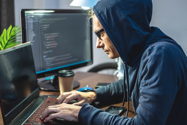 Haker w kapturze programuje kod wirusa na klawiaturze laptopa