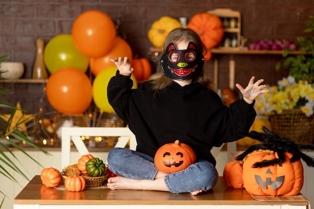 Halloween, dziecko dziewczynka w stroju wiedźmy i karnawałowej masce w ciemnej kuchni z dyniami i wielkim pająkiem straszy rękoma, uśmiecha się, raduje