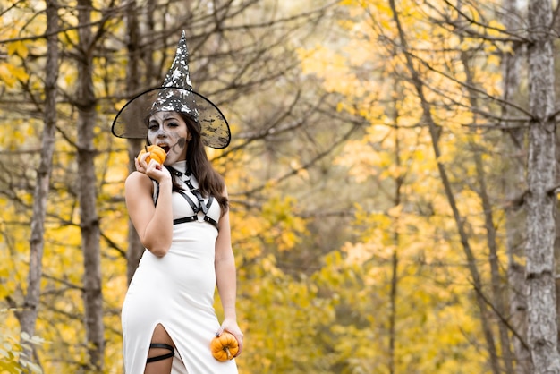 Halloween Młoda piękna dziewczyna w białej sukni z makijażem na twarzy w kapeluszu wiedźmy pozuje w jesiennym lesie Makijaż twarzy szkieletu Gryzie małą dynię Dzień zmarłych