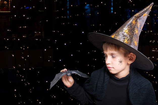 Halloweenowe dzieciaki. Chłopiec w kapeluszu czarodzieja iz namalowanym pająkiem na policzku, trzymający czarnego nietoperza Halloween, by straszyć kogoś na czarnym tle z bokeh. Gotowy na świąteczny smakołyk.