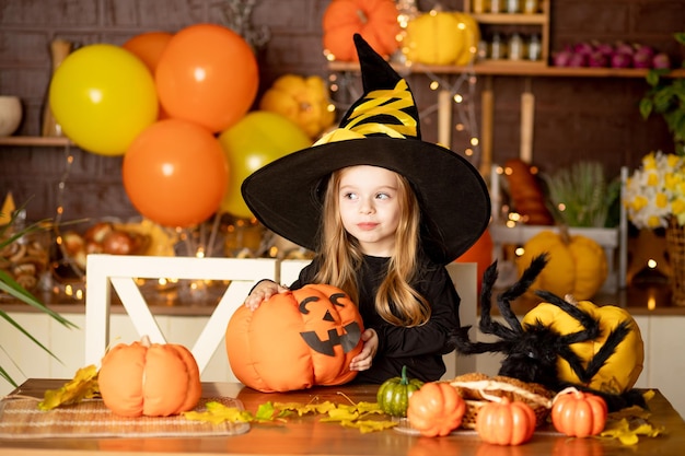 Halloweenowe dziecko dziewczynka w stroju wiedźmy z dyniami i dużym pająkiem w ciemnej kuchni dekoruje dynię podczas obchodów Halloween