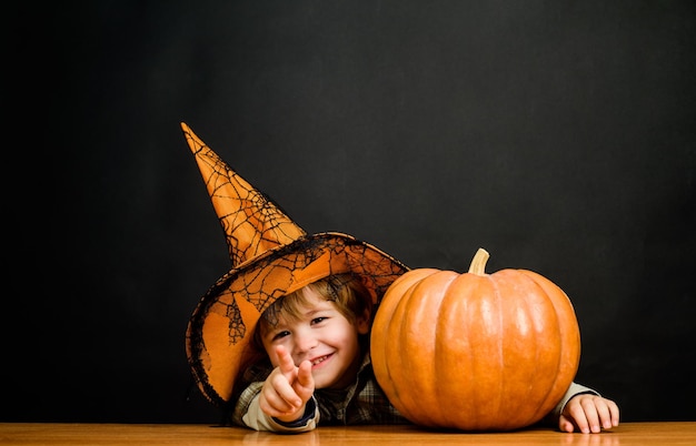 Halloweenowy chłopiec wskazujący do przodu. Dziecko z dynią, wskazując na ciebie. Mały chłopiec w kapeluszu wiedźmy z halloweenową dynią pokazuje palec na tobie. Cukierek albo psikus. Przygotowanie święta Halloween. Impreza halloween'owa.