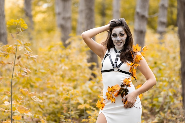 Halloweenowy portret młodej pięknej dziewczyny w białej sukni z makijażem na twarzy Pozowanie w jesiennym lesie Makijaż twarzy szkieletu Dzień Zmarłych