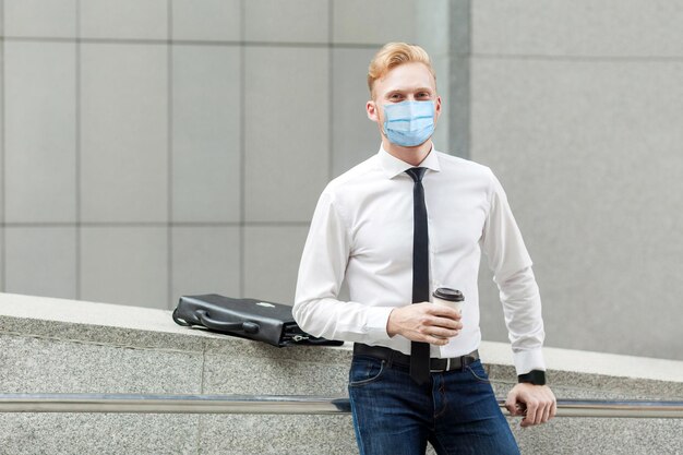 Hamulec na kawę młody przystojny biznesmen z chirurgiczną maską medyczną trzyma kubek patrząc na kamerę