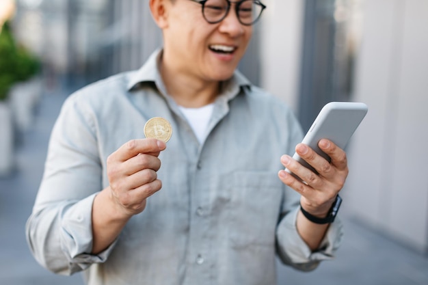 Handel online Szczęśliwy azjatycki mężczyzna trzymający złote bitcoiny i smartfon w średnim wieku, zarabiający kryptowalutę