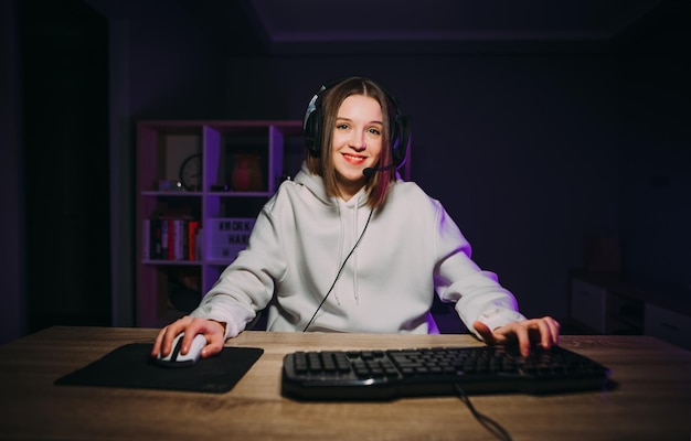 Happy girl gamer w zestawie słuchawkowym siedzi w domu przy komputerze i z uśmiechem gra w gry wideo