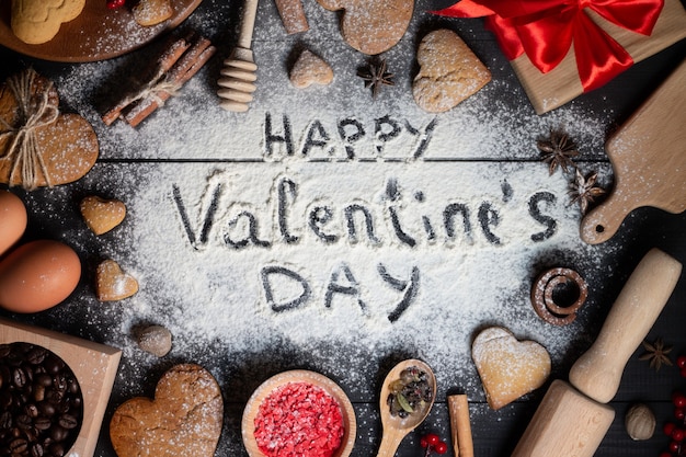 Happy Valentines Day napisane na mące. Piernikowe ciasteczka w kształcie serca, przyprawy, ziarna kawy i artykuły do pieczenia