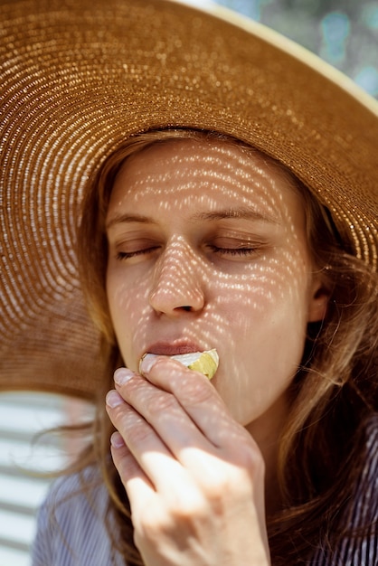 Headshot kobiety w słomkowym kapeluszu lato jedzenie grillowanych warzyw na zewnątrz, oczy zamknięte. Wzór światła i cienia na twarzy