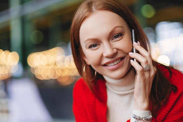 Heashot uroczej europejskiej przedsiębiorczyni ma zdrową skórę przyjemny uśmiech ubrana w czerwone ubrania patrzy prosto na aparat trzyma nowoczesny telefon komórkowy poza rozmytym tłem