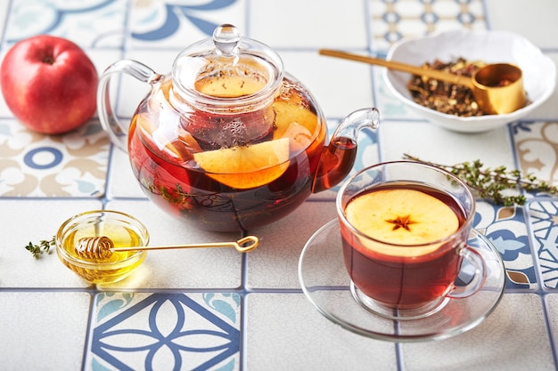 Herbata owocowa z jabłkami i tymiankiem w szklanym czajniczku i filiżance na stole z kolorowych płytek