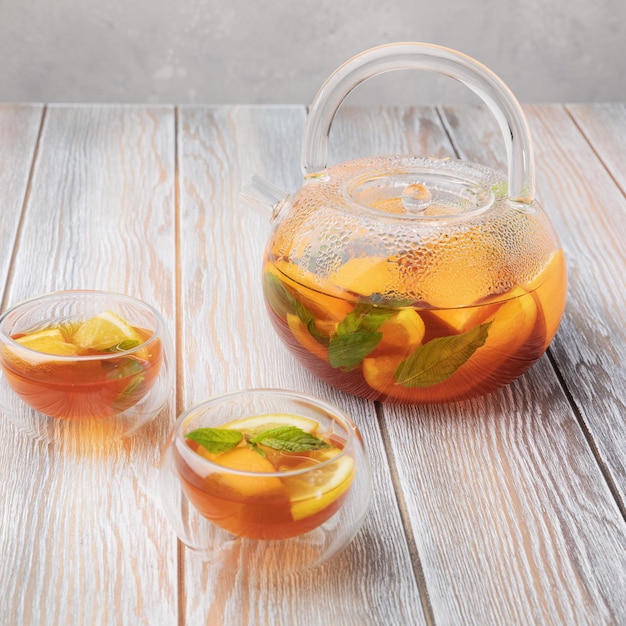 Zdjęcie herbata owocowa z pomarańczą, cytryną i miętą w szklanym czajniczku i dwie filiżanki (miski) na drewnianym tle
