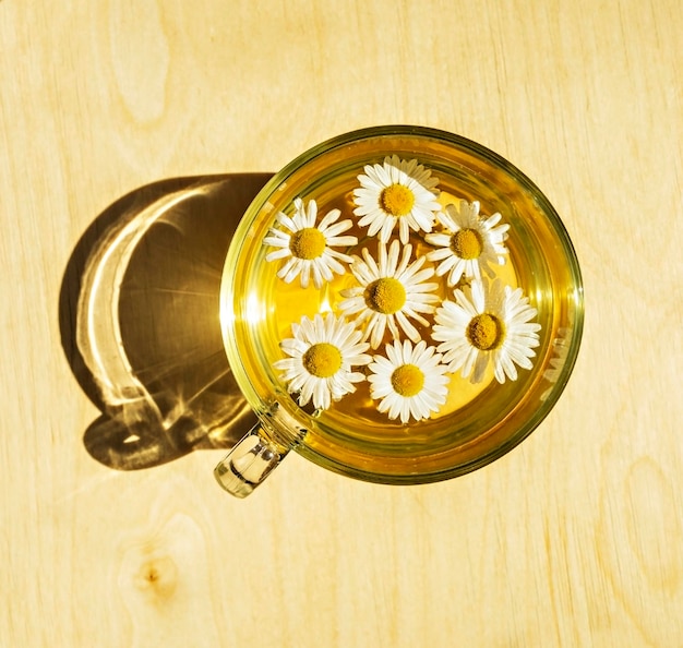 Herbata rumiankowa w szklanym kubku z kwiatami rumianku na jasnym drewnianym tle