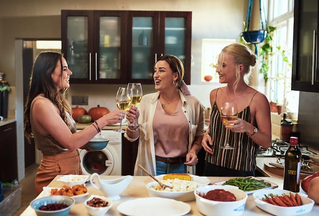 Heres to us girls Przycięte zdjęcie grupy wesołych młodych przyjaciół wznoszących uroczysty toast z napojami, stojąc w kuchni przygotowującej jedzenie w domu