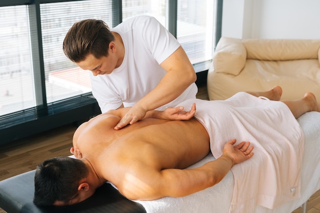 Highangle widok profesjonalnego masażysty z silnymi rękami masuje łopatki i tył muskularnego sportowca leżącego na brzuchu przy stole do masażu w salonie Koncepcja zdrowego stylu życia