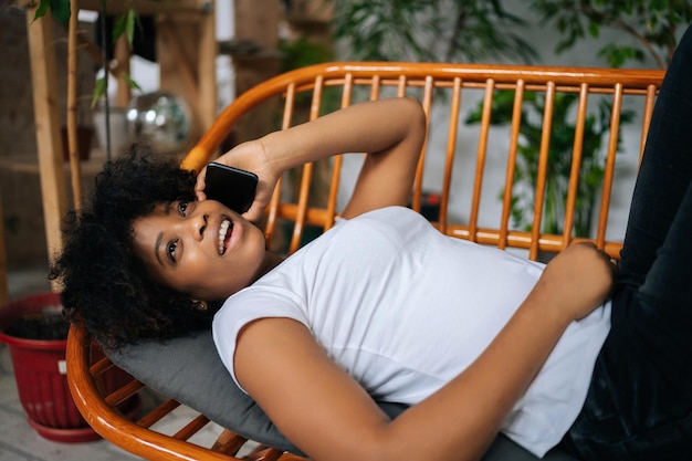 Highangle widok szczęśliwej uroczej czarnej kobiety rozmawiającej na smartfonie, leżącej na wygodnej kanapie, mającej przyjemną rozmowę telefoniczną w salonie w domu