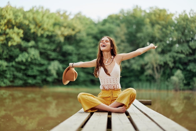 Hippie ekoaktywistka podróżująca siedzi na moście nad jeziorem z wyciągniętymi ramionami z kapeluszem i szczerze uśmiechając się Zdjęcie wysokiej jakości