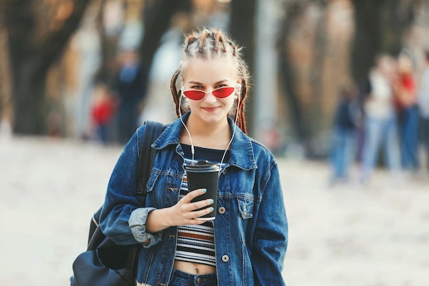 Hipster atrakcyjna dziewczyna z dredami w słuchawkach i okularach ubrana w dżinsową kurtkę, pijąca kawę i chodząca po ulicy