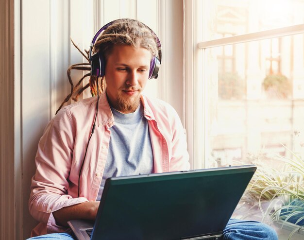 Hipster atrakcyjny brodaty mężczyzna z dredami w słuchawkach siedzący przy oknie i rozmawiający w sieciach społecznościowych