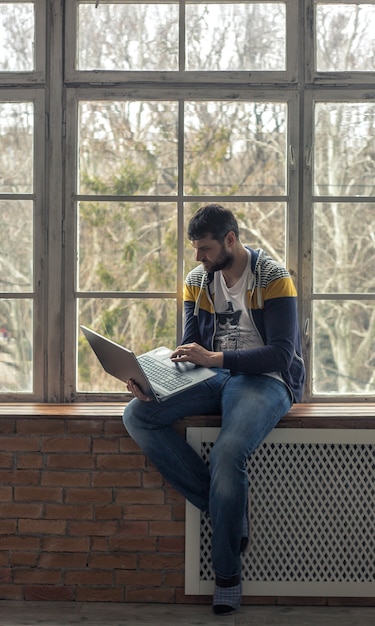 Hipster brodaty mężczyzna w kapciach przy laptopie lub notebooku siedząc na parapecie