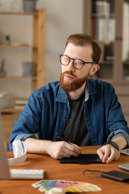 Hipster Brodaty Mężczyzna W Okularach Siedzi Przy Stole I Używa Tabletu Digitizera, Myśląc O Wizualnym Rozwiązaniu