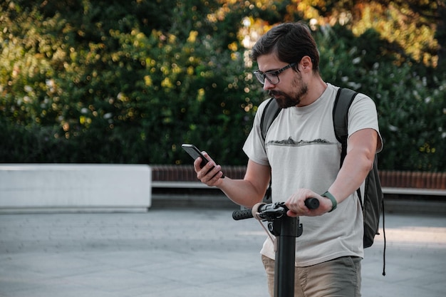 Hipster człowiek dojeżdżający do pracy ze skuterem elektrycznym na zewnątrz w mieście za pomocą smartfona