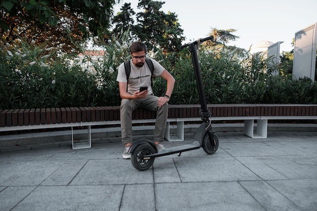 Hipster człowiek dojeżdżający do pracy ze skuterem elektrycznym na zewnątrz w mieście za pomocą smartfona