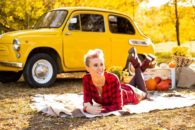 Hipster kobieta z krótkimi włosami leżąca na kocu w tle jesiennego parku w pobliżu żółtych kwiatów samochodu i dyni