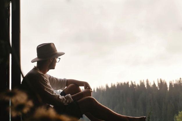 Hipster mężczyzna siedzi na ganku drewnianego domu patrząc na las w wieczornym spokojnym momencie letnich wakacji koncepcja miejsca na tekst