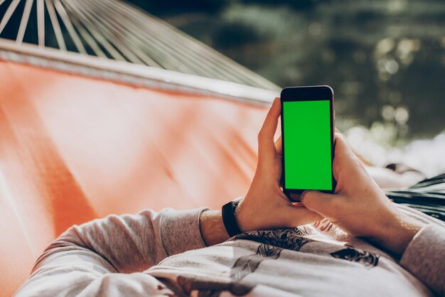Hipster mężczyzna trzyma inteligentny telefon z pustym ekranem z miejscem na tekst i relaks w hamaku w słonecznym letnim parku makieta faceta patrzącego na pusty telefon koncepcja mediów społecznościowych