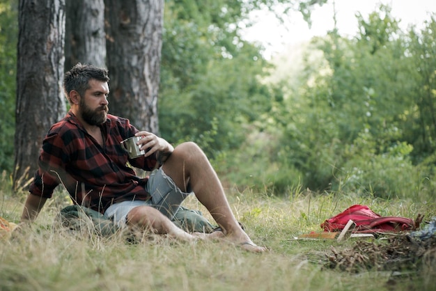 Hipster pić herbatę lub kawę przy ognisku Brodaty mężczyzna zrelaksować się przy kubku przy ognisku Facet cieszyć się kempingiem w lesie Koncepcja letnich wakacji Kemping i turystyka i podróże