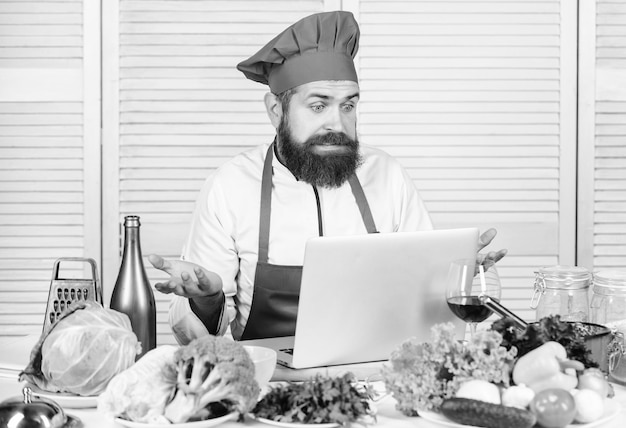 Hipster w kapeluszu i fartuchu uczy się gotować online Edukacja kulinarna online Koncepcja e-learningu Mężczyzna szef kuchni szuka przepisu internetowego gotowanie żywności Laptop szefa kuchni czyta przepisy kulinarne Szkoła kulinarna
