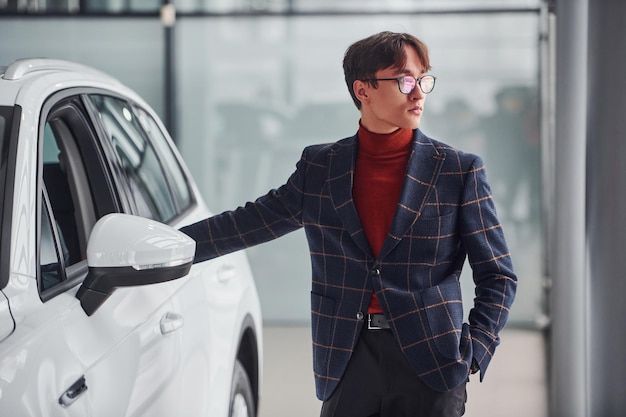 Hipster w wizytowym i w okularach stojący w pobliżu białego nowoczesnego samochodu w pomieszczeniu.