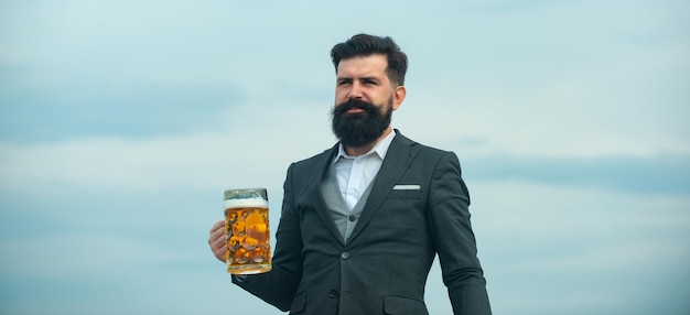 Hipster z brodą i wąsami w garniturze pije piwo. Czas na piwo. Przystojny barman trzyma szklankę piwa na niebie. Baner, miejsce na kopię.