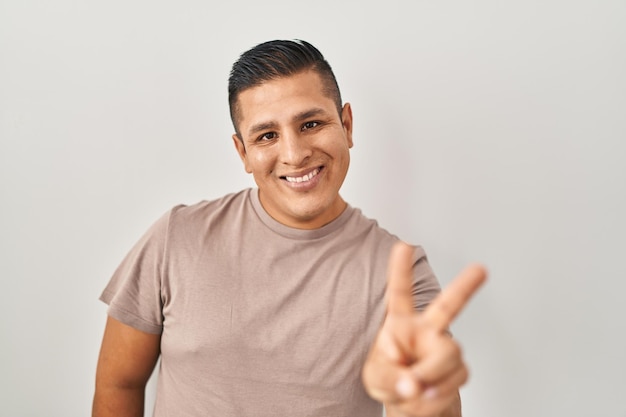Hiszpański młody człowiek stojący na białym tle uśmiechający się, patrzący w kamerę, pokazujący palce temu znakowi zwycięstwa numer dwa