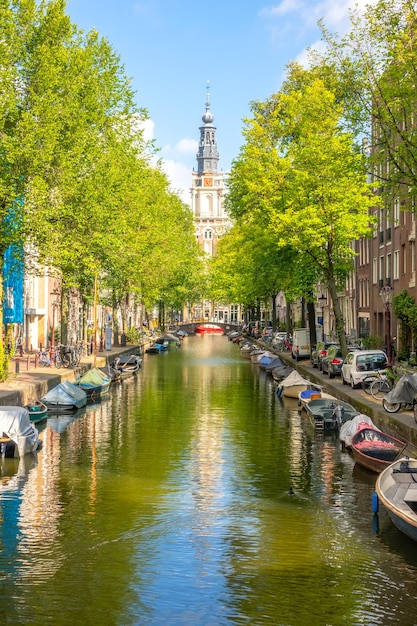 Holandia Słoneczny letni dzień nad kanałem Amsterdam Zaparkowane samochody na nabrzeżu i wiele zacumowanych łodzi na wodzie Budynek katedry w oddali