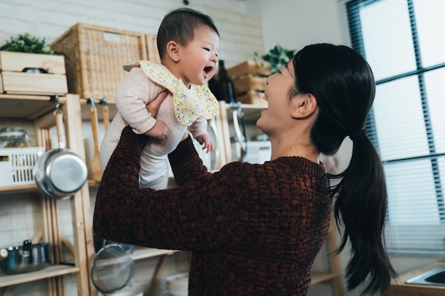 holenderski kąt strzału radosny asian nowa matka uśmiecha się i podnosząc jej słodkie dziecko podczas zabawy z nią w jasnej kuchni w domu.