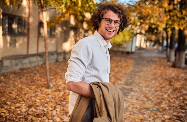 Horyzontalny portret młodego biznesmena w okularach pozuje na zewnątrz idąc na lunch Student na jesiennej ulicy Inteligentny facet ubrany na co dzień nosi okulary z kręconymi włosami idąc ulicą