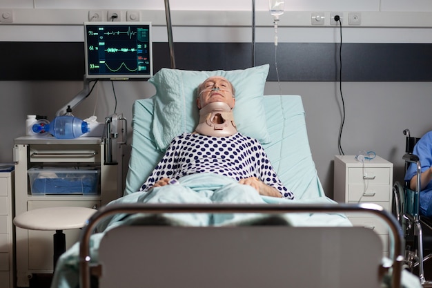 Zdjęcie hospitalizowany senior leżący nieprzytomny w szpitalnym łóżku z kołnierzem ortopedycznym, mający poważne...