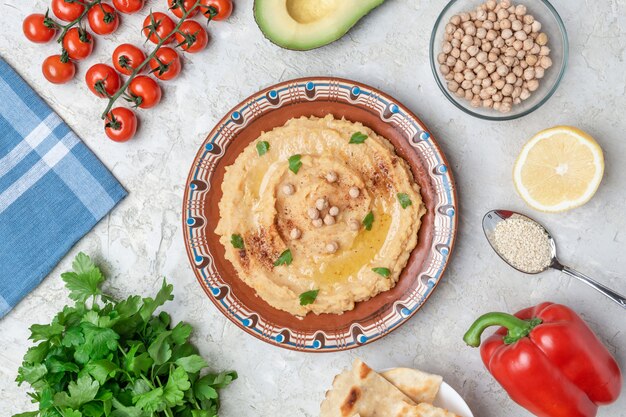 Hummus do brązowej płytki glinianej z niebieskim wzorem. Na białym stole znajdują się warzywa, warzywa, trójkątne kawałki pity. Widok z góry. Leżał płasko.