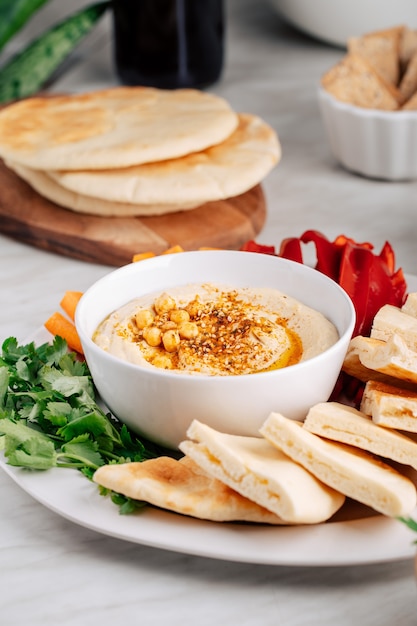 Hummus w misce i chleb pita na jasnym tle z warzywami.