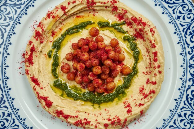 Hummus z czerwonymi przyprawami, ciecierzycą i ziołami na ceramicznym talerzu na drewnianej powierzchni. Kuchnia arabska lub izraelska. Z bliska, poziome. Przekąska