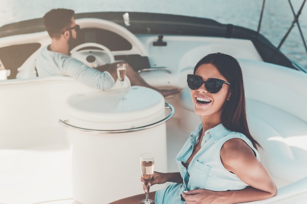 Idealny czas razem. Piękna młoda kobieta trzymająca kieliszek z szampanem i patrząca w kamerę, podczas gdy jej chłopak prowadzi jacht